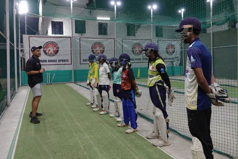 Coach briefing a line of helmeted batters inside the lit Harvard House Sports indoor nets